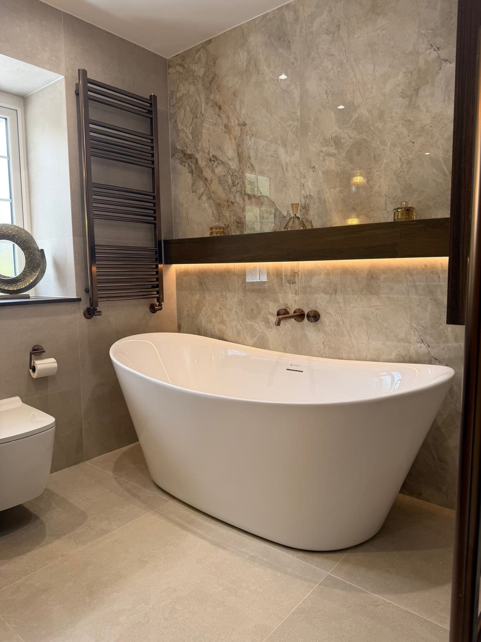 A freestanding bath beneath a softly lit timber shelf, by Holden & Salter